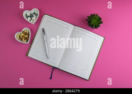 An open notebook with a silver pen, next to pills and nuts, everything stands on a pink background. Flat lay Stock Photo