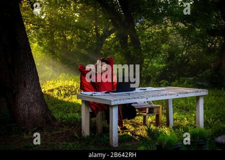 Girl works at a laptop in the garden in the summer. The work of a freelancer. Working remotely. Early morning outdoors Stock Photo