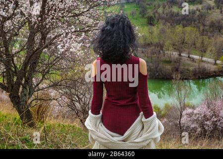 Girl outdoors on a background of a spring landscape with blooming trees and a lake. A view from the back of a girl in a burgundy dress and white sweat Stock Photo