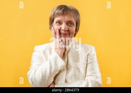 Portrait of a smiling gray-haired senior woman with palm of hand holding cheek on yellow background.