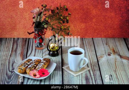 Warm autumn still life, a plate with eclairs, a cup of coffee, an old-fashioned lamp and a vase with a herbarium on a wooden table. Close-up. Stock Photo