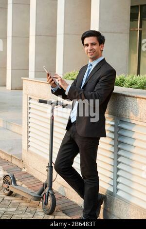 Full-length portrait of smiling young businessman using application on tablet computer when standing outdoors next to his electric scooter Stock Photo