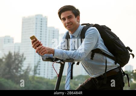 Portrait of handsome young smiling businessman with big backpack holding smartphone and riding on electric scooter Stock Photo