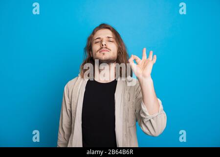 Long haired caucasian man with beard is gesturing the okay sign while posing confidently on a blue background Stock Photo