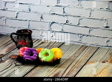 A rectangular plate with five multi-colored donuts and a black ceramic coffee mug stand on a wooden table. Close-up. Stock Photo