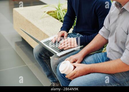 Cropped image of software developer typing on laptop when talking to his mature coworker during coffee break Stock Photo