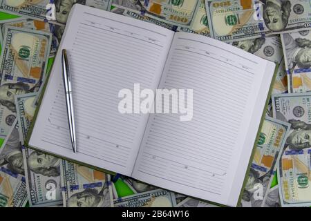 An open notebook with a pen stand on a pile of dolar banknotes Stock Photo