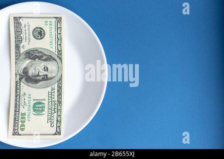 Medicines costs money. Drugs and dollars.White plate with multi-colored pills and capsules and a 100 dollar bill on a classic blue background. Pills Stock Photo