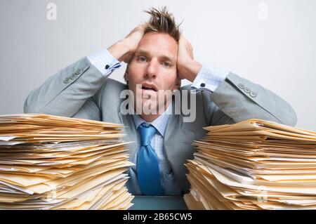 Fatigued office worker holding his head in his hands on the piles of file folders on his desk Stock Photo