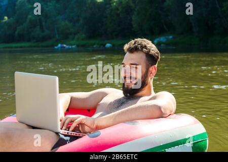Young man with laptop on inflatable ring in water. attractive smiling bearded guy looks at laptop screen. Workaholic, work on vacation Stock Photo