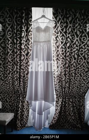 White Wedding dress hang on a shoulders near window, before ceremony Stock Photo