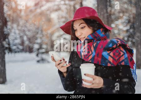 Fashionably dressed woman in an overcoat and wide-brimmed hat in winter Park drinking coffee and talking on the smartphone Stock Photo