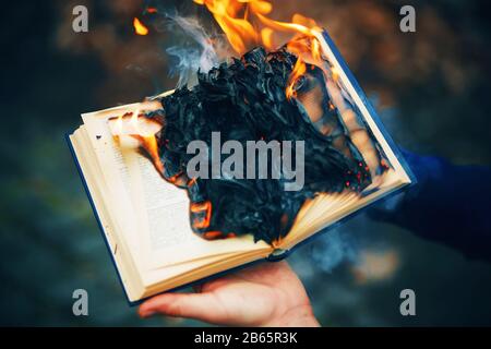 A man holds in his hands a thick old book, the pages of which burn with a bright red flame and turn into black ash. Stock Photo
