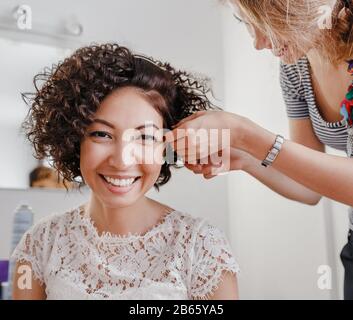 Professional hairdresser artist making curly hairstyle to charming Brunette bride woman with short hair on her wedding day. Beauty Studio interrior Stock Photo