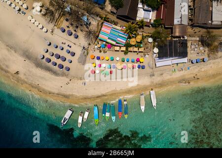 Top down aerial view of colorful boats and sunshades on a tropical beach on a small island fringed by a coral reef