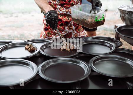 The black-gloved cook puts the salad on a black, round plate. Picnic. Stock Photo