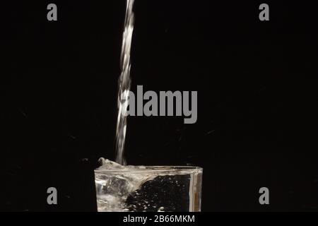 Water jet falling into a glass and generating a splash of water droplets splashing everywhere. Transparent water drops on black background. Stock Photo