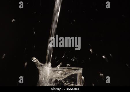 Water jet falling into a glass and generating a splash of water droplets splashing everywhere. Transparent water drops on black background. Stock Photo