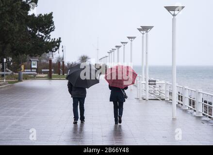 10 March 2020, Schleswig-Holstein, Lübeck-Travemünde: Two walkers with umbrellas are walking along the almost deserted promenade on the beach by the water of the Baltic Sea. Photo: Daniel Bockwoldt/dpa Stock Photo