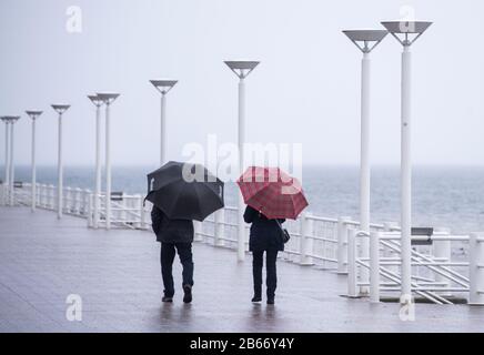 10 March 2020, Schleswig-Holstein, Lübeck-Travemünde: Two walkers with umbrellas are walking along the almost deserted promenade on the beach by the water of the Baltic Sea. Photo: Daniel Bockwoldt/dpa Stock Photo