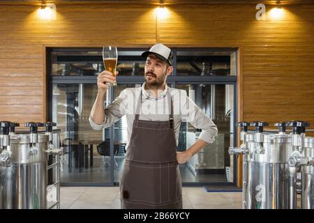 Professional brewer on his own craft alcohol production. Specialist, man wearing in workwear testing, trying product on its taste, color and smell. Concept of open business, eco product, craft brewery. Stock Photo