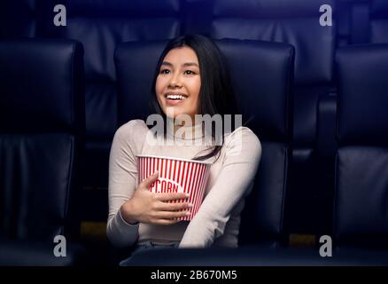 Going to movies alone concept. Pretty Asian woman with popcorn enjoying film in cinema Stock Photo