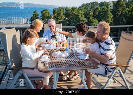 Big happy family having breakfast outdoors on terrace together, sitting around table, drinking coffee. Beautiful sea view, warm summer morning. Family Stock Photo