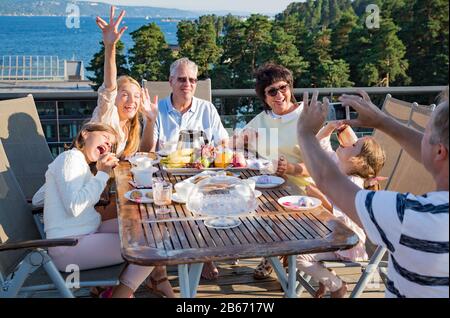 Big happy family having breakfast outdoors on terrace together, sitting around table, drinking coffee. Father taking selfie on phone. Beautiful view Stock Photo