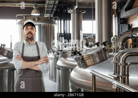 Professional brewer on his own craft alcohol production. Specialist, man wearing in workwear posing confident with hands crossed. Concept of open business, eco product, craft brewery, individual factory. Stock Photo