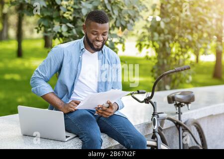Happy Black Man With Laptop And Bike Checking Business Documentation Outdoors Stock Photo