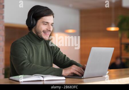 Young positive man in wireless headphones studying online on laptop Stock Photo