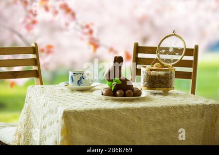 Beautiful served small table with Easter decorations on outdoor terrace. Little chocolate bunny with bow, chocolate eggs, cup of coffee, crystal bowl. Stock Photo