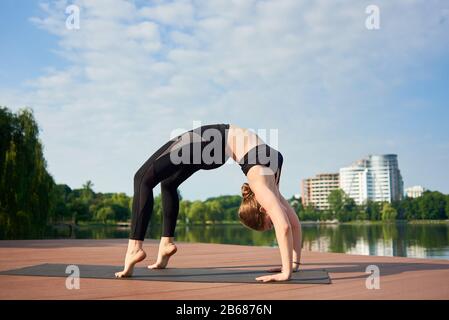 Slim girl in black sportswear standing in pose of bridge, asana urdhva dhanurasana in a city park with green trees, lake and buildings in background under blue sky with copy space. Morning training Stock Photo