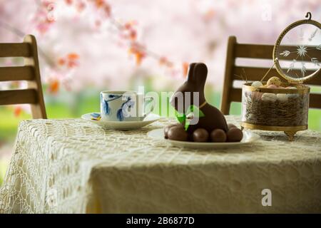 Beautiful served small table with Easter decorations on outdoor terrace. Little chocolate bunny with bow, chocolate eggs, cup of coffee, crystal bowl. Stock Photo