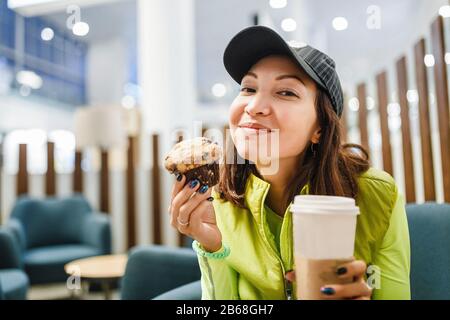 Young brunette woman drinking coffee and eating a delicious muffin cake in modern airport cafe, fastfood travel concept Stock Photo