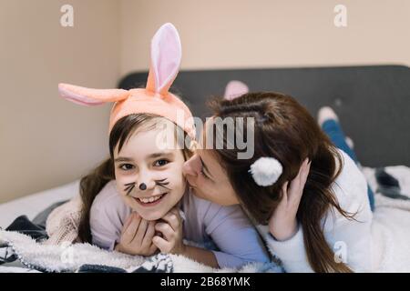 Mother kisses her little daughter with bunny ears. Cute child girl smiling and wearing bunny ears for Easter while mother kissing her. Stock Photo