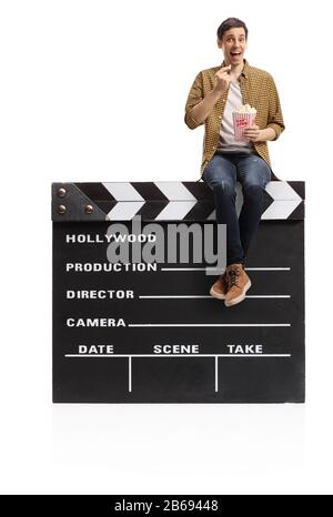 Young man sitting on a big movie clapper board and eating popcorn isolated on white background Stock Photo