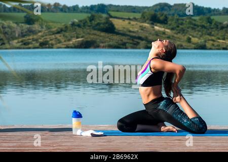 Young attractive smiling woman practicing yoga, sitting in One Legged King Pigeon exercise, Eka Pada Rajakapotasana pose.Morning workout against the b Stock Photo