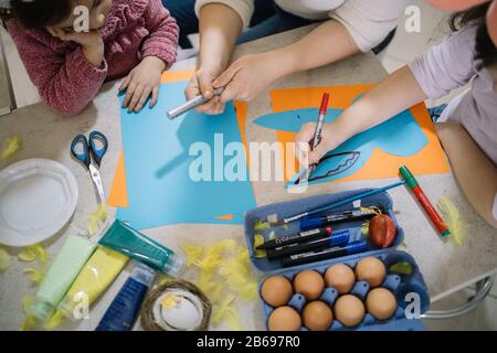 Children's hands drawing on paper with felt tipped pen. Top view of girls at table with eggs, paint, feathers, brushes, markers,scissors and paper she Stock Photo
