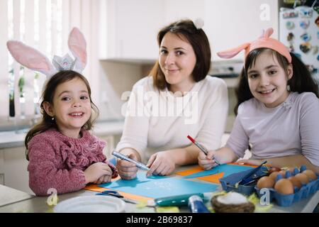 Happy woman and girls in bunny ears painting. Smiling mom and kids wearing rabbit ears painting and drawing together while looking at camera. Stock Photo