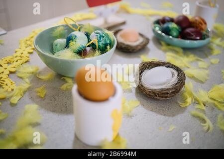 Painted Easter eggs with feathers and nest. Out of focus egg on ceramic bunny stand on kitchen table with with Easter decoration, nests, painted eggs Stock Photo