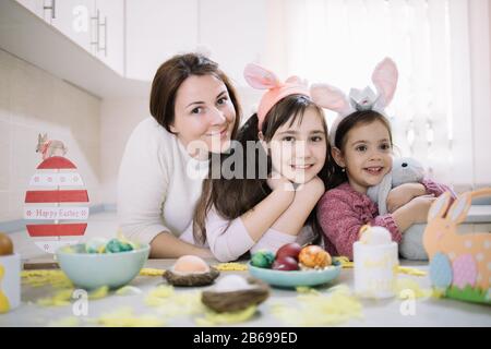Smiling mother and daughters in decorated for Easter kitchen. Happy family of mom and two sisters wearing rabbit ears and one of them hugging plush bu Stock Photo