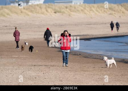 Woman in red coat talking on mobile phone while walking her dog among other dog walkers on Blyth Beach, Northumberland Stock Photo