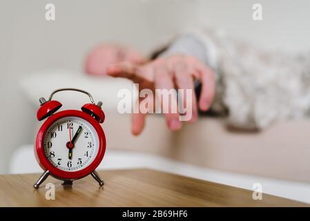 Red old style alarm clock with sleeping man on background. Man holding his hand to clock, wants to switch it off Stock Photo