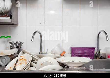 Large metal sink with dirty dishes in professional restaurant kitchen, stack of messy white plates, crockery, appliances with leftover food, water tap Stock Photo