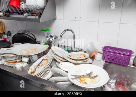 Large metal sink with dirty dishes in professional restaurant kitchen, stack of unclean white plates, crockery, cutlery with leftover food, water tap, Stock Photo