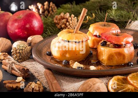 Baked apples on brown plate with holiday Christmas decoration with Christmas tree branch, cones, cinnamon, walnuts and red apple. Stock Photo