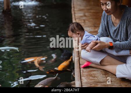 asian mother and her baby daughter see koi fish from the edge of a pond Stock Photo