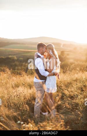 Man and woman in stylish boho casual clothes, posing on the background of mountains, field at sunset. Stylish hipster couple hugging Stock Photo