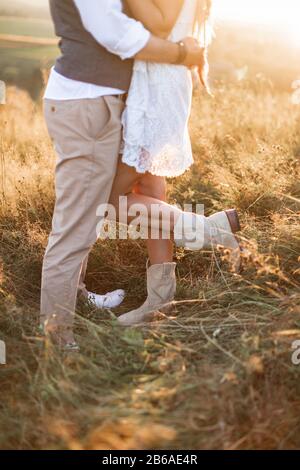 Cropped image of legs of stylish rustic couple, boho woman in white dress and cowboy boots and man in shirt and pants posing in sunny field, hugging